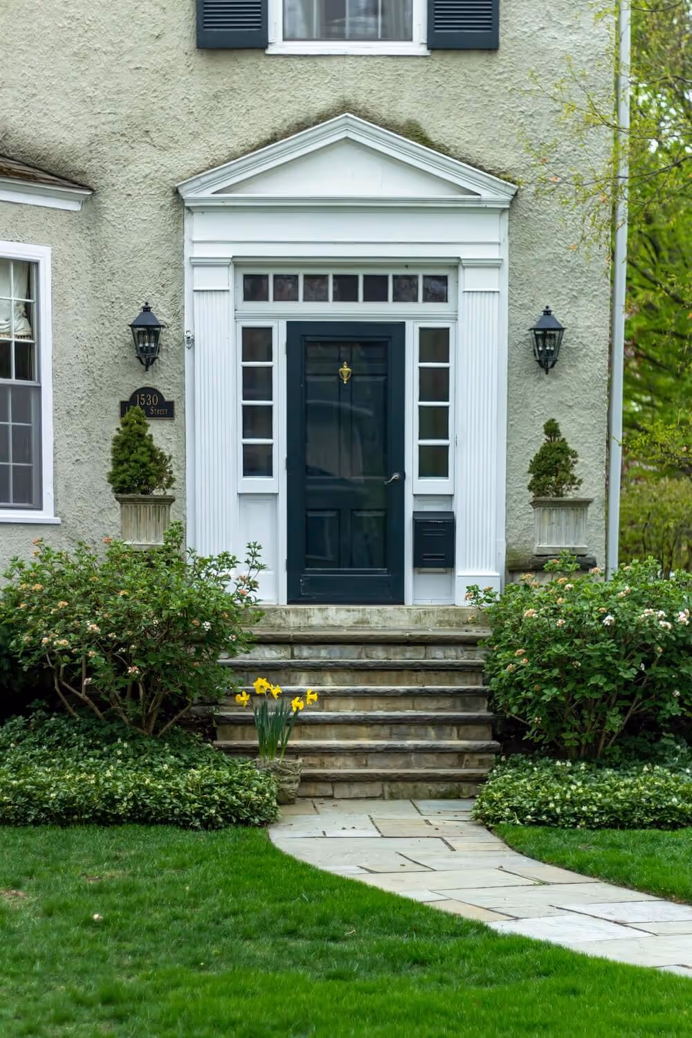 Front entrance of a house with a black door framed by white columns and windows, stone steps, green bushes, and yellow flowers.