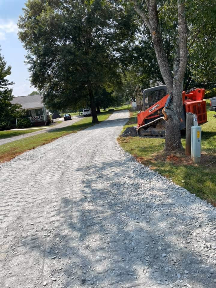 Gravel driveway with a Kubota construction vehicle parked near a tree on a sunny day in a residential area.
