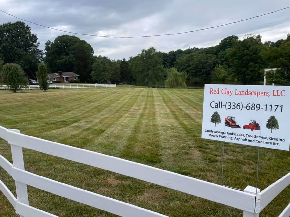 Large mowed lawn with striped grass pattern, white fence in foreground, and a sign for Red Clay Landscapers offering various landscaping services and phone number.