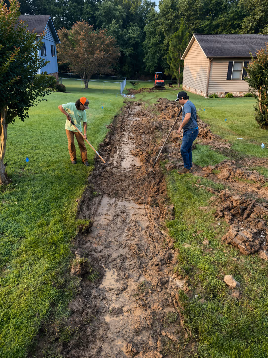 Two men working with shovels in a muddy trench between green lawns near houses.