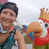 Smiling woman wearing a cap and turquoise sportswear posing next to a colorful bird statue with a crown outdoors.
