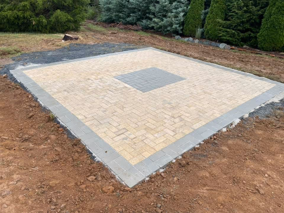Square outdoor patio area made with beige and gray paving stones, surrounded by dirt and greenery.