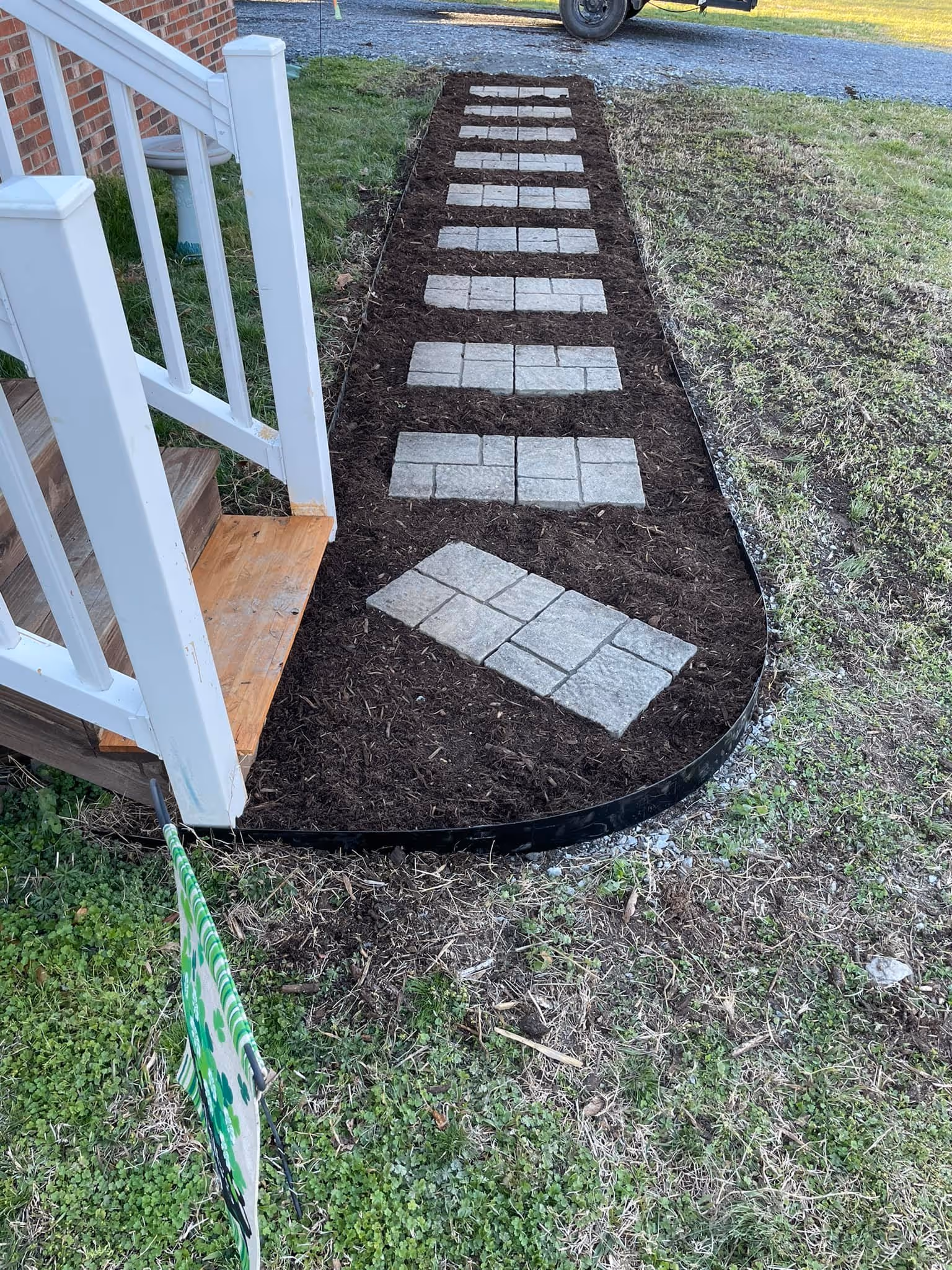 Stone pathway with rectangular stepping stones embedded in dark mulch, bordered by grass and leading from a wooden porch with white railings.