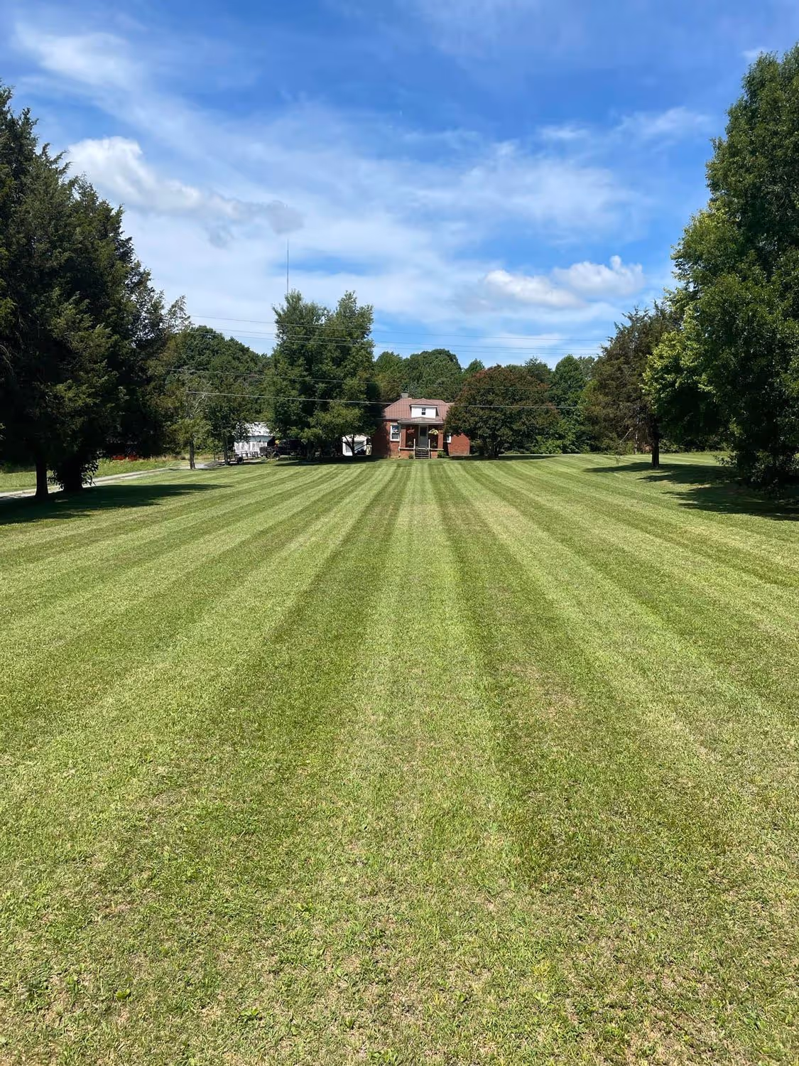 Wide, neatly mowed lawn with striped grass leading to a small brick house surrounded by trees under a partly cloudy blue sky.
