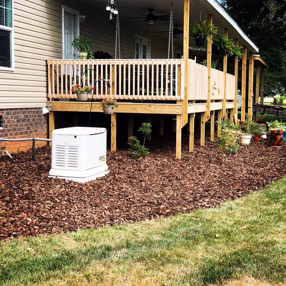 Wooden raised deck with hanging plants and fresh mulch around its base next to a beige house with brick foundation.